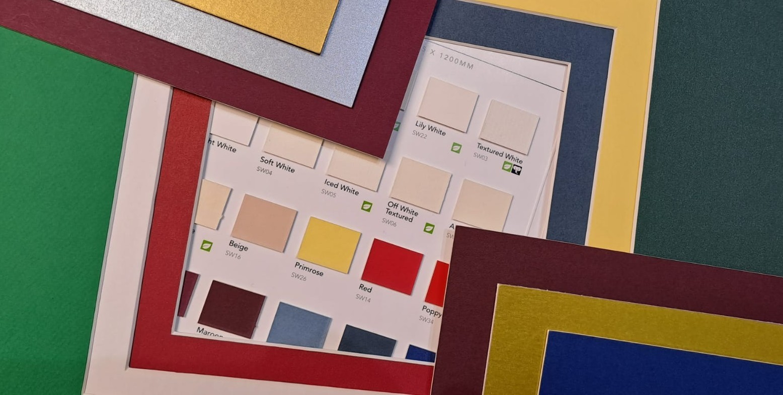 Several colourful photo mount boards are layered on top of each other, partially revealing a colour sample sheet with various shades and names for selection in the background.